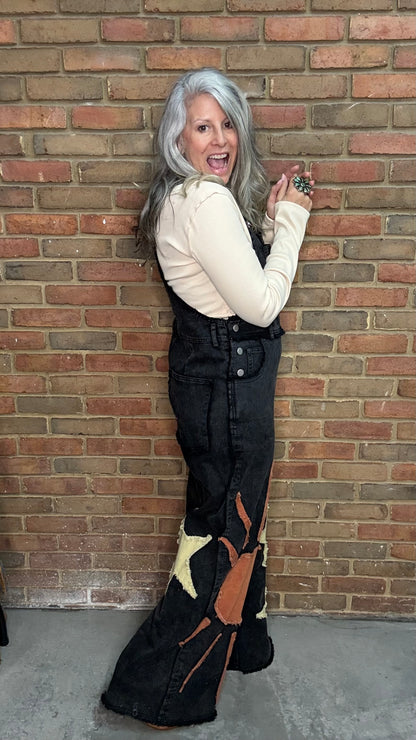 A woman with long gray hair poses in front of a brick wall wearing a cream long-sleeve top and black denim overall flare pants with a brown and beige abstract graphic print near the hem.