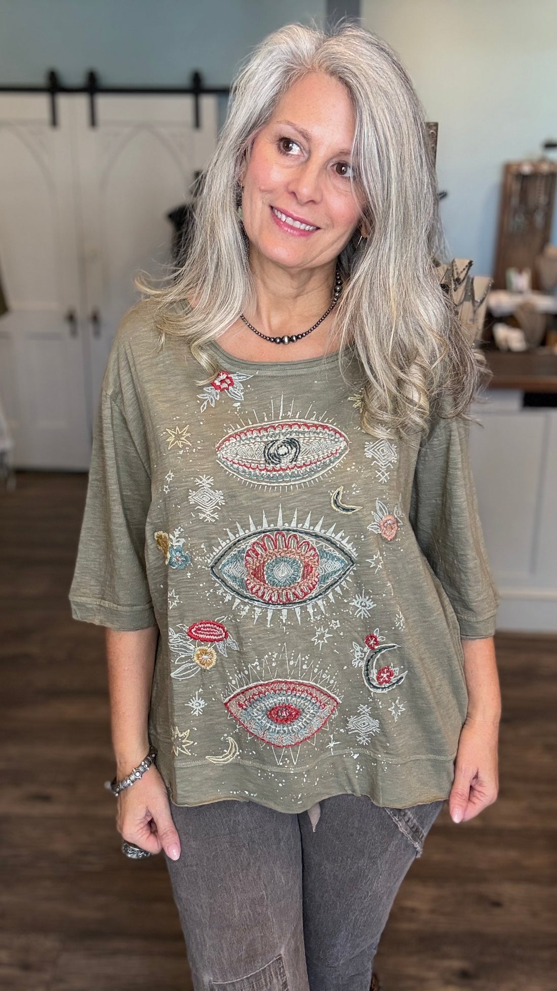 A woman with long gray hair wearing a sage green tunic top with colorful embroidered eyes, stars, and moons, paired with gray jeans in an indoor setting.