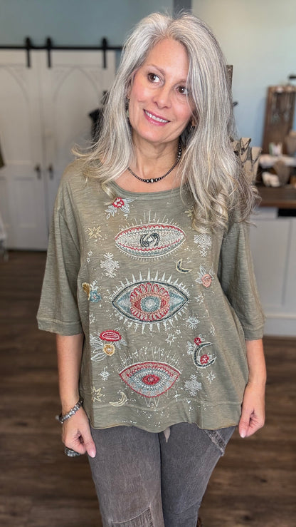 A woman with long gray hair wearing a sage green tunic top with colorful embroidered eyes, stars, and moons, paired with gray jeans in an indoor setting.
