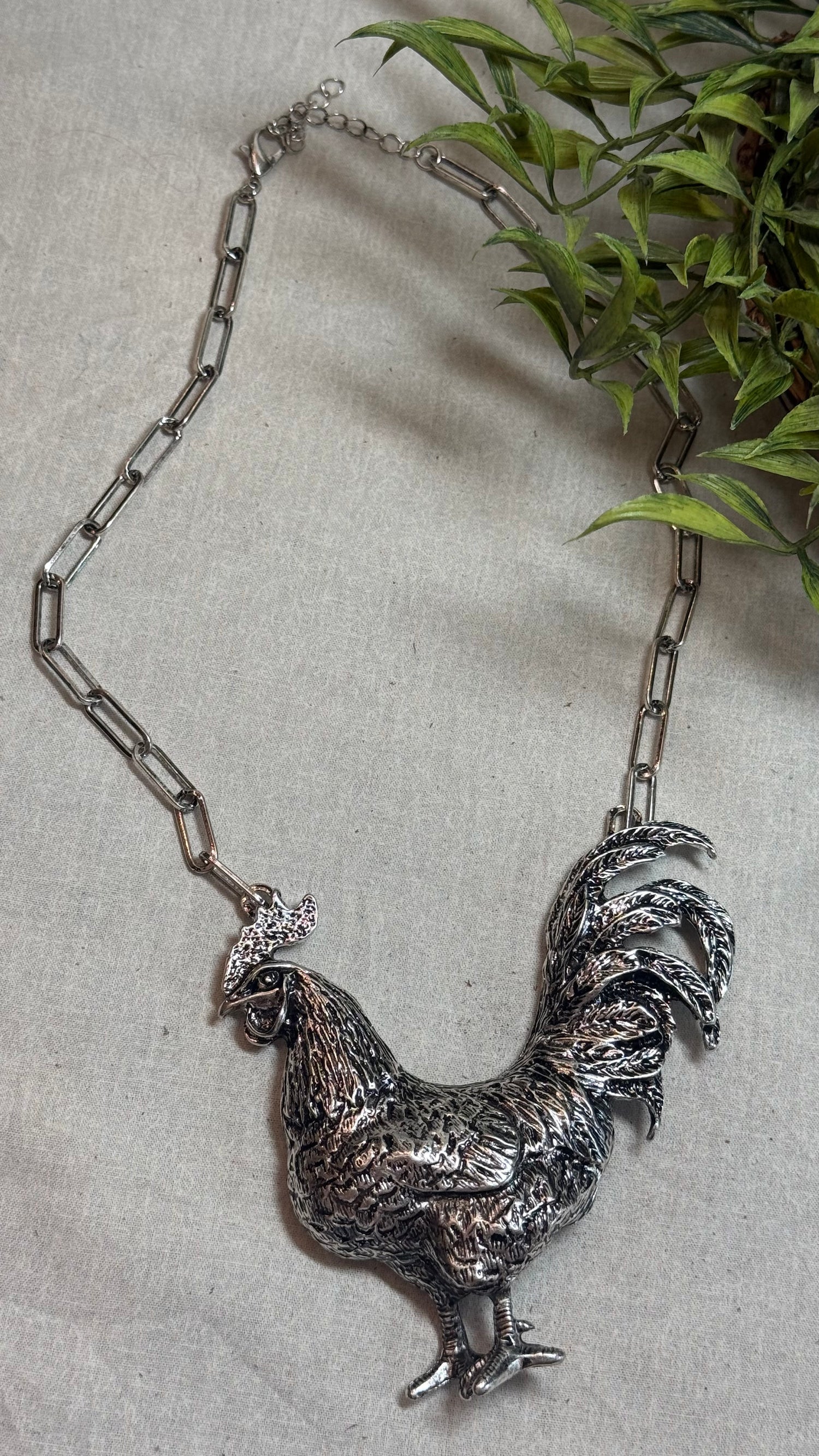 A detailed silver rooster pendant necklace on a silver link chain, displayed on a textured light gray fabric background with green leaves in the upper right corner.