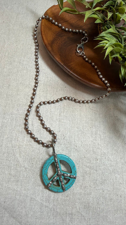 A beaded necklace with a turquoise peace sign pendant, styled on a light beige linen fabric next to a wooden dish and green leaves.