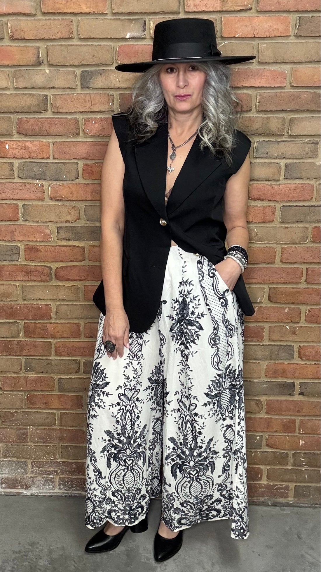 Person wearing a black vest, Floral Print Wide Leg Pant, and a black hat against a brick wall.