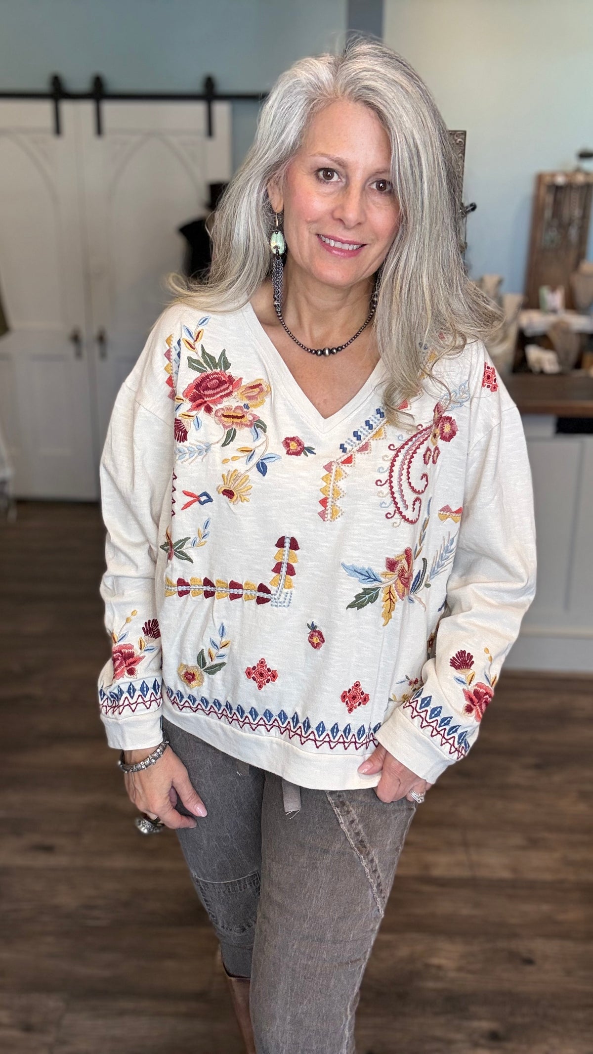 A woman with long gray hair wearing a cream-colored long-sleeve V-neck top with colorful floral and paisley embroidery, paired with gray pants, standing in a rustic indoor setting.