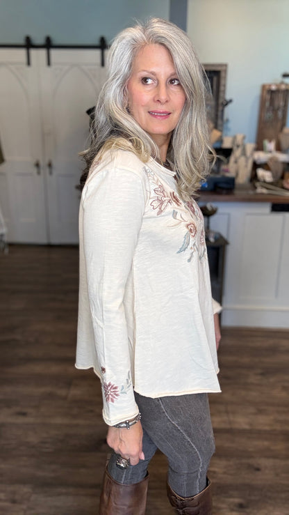 A woman with long gray hair wearing a cream-colored long-sleeve top with floral embroidery on the chest and cuffs, paired with gray pants and brown boots, standing in a home interior setting.