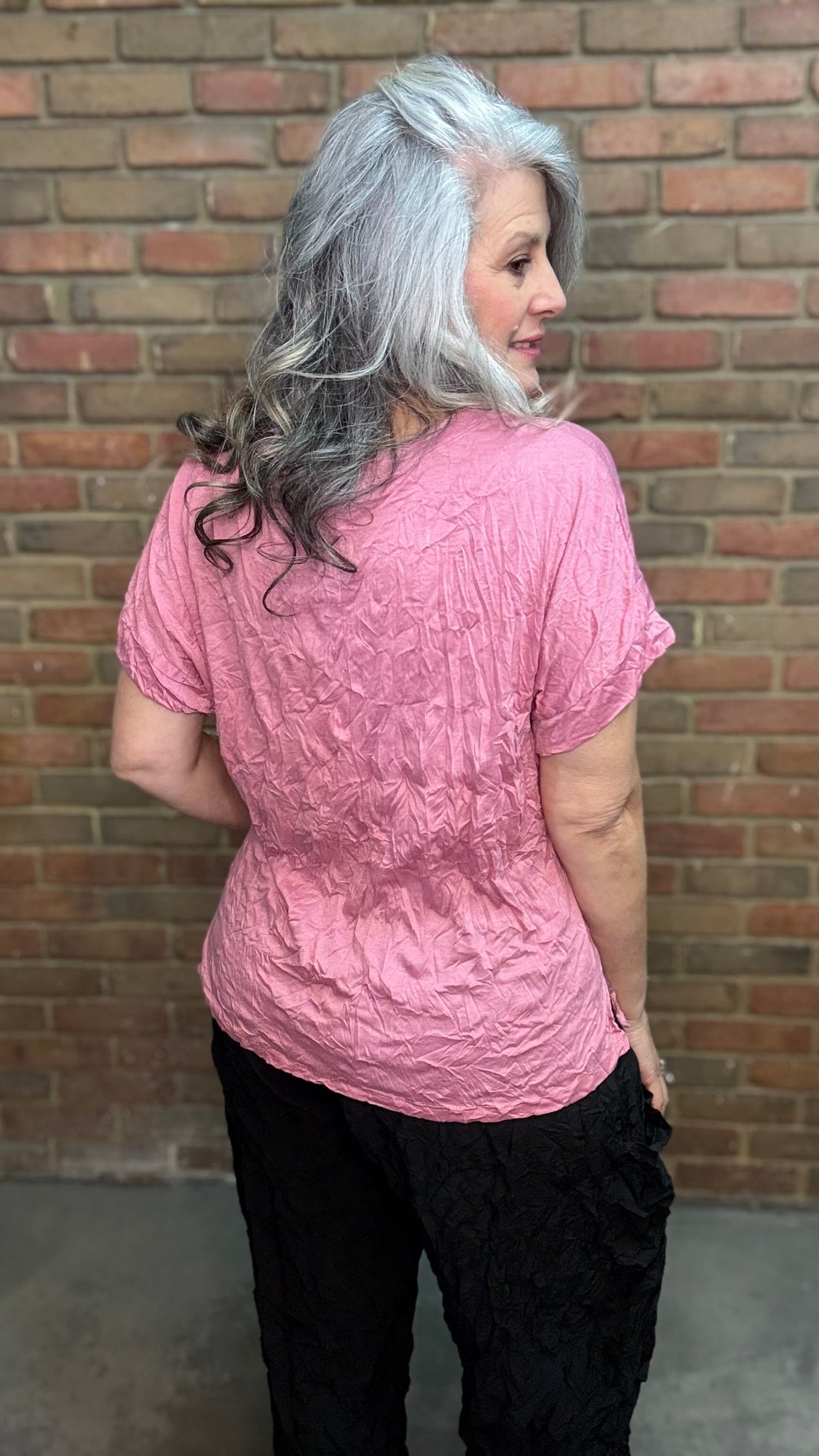 Woman with gray hair wearing Rose Dolman Sleeve Top and black pants standing in front of a brick wall.