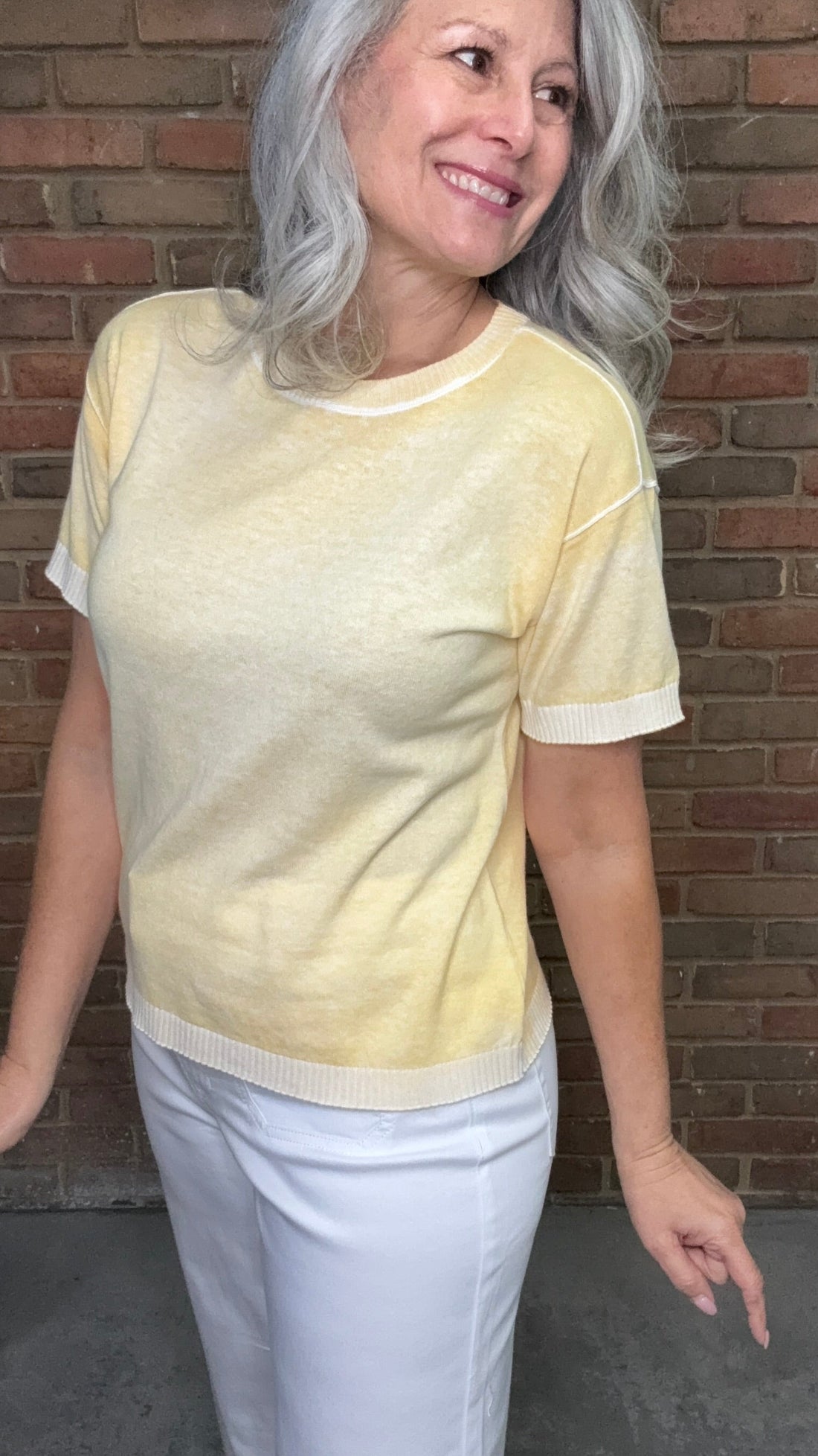 Woman wearing the Sunshine Short Sleeve Sweater, a light yellow sweater and white pants standing against a brick wall.