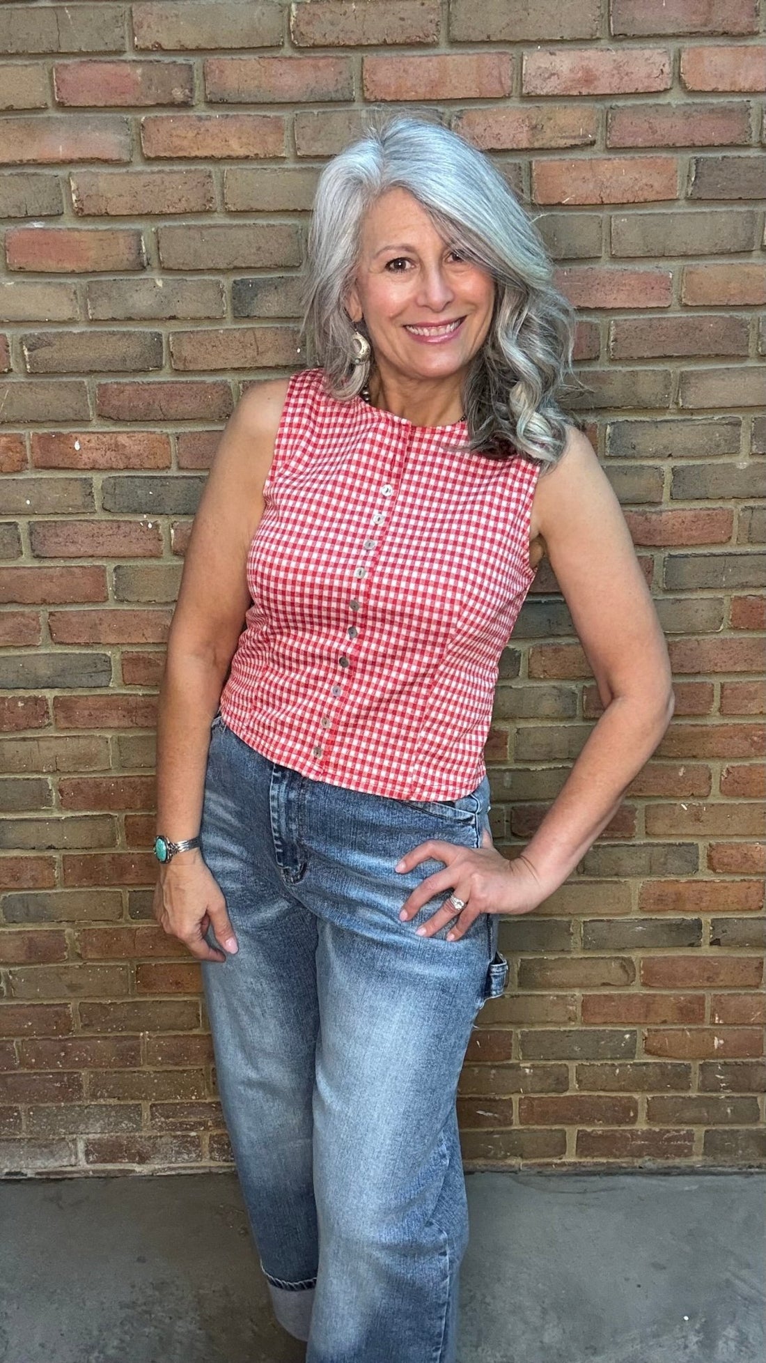 Woman wearing the Red Plaid Sleeveless Shirt  and blue jeans standing against a brick wall.