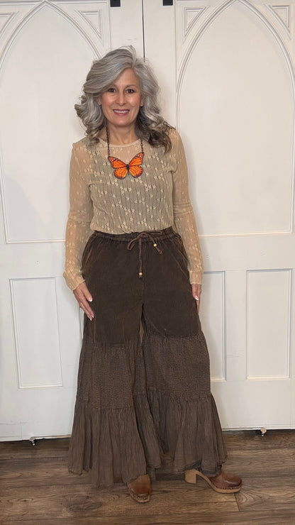 Woman wearing a beige lace top with Monarch Butterfly Necklace and Amber Boho Chic pants against a white paneled wall.