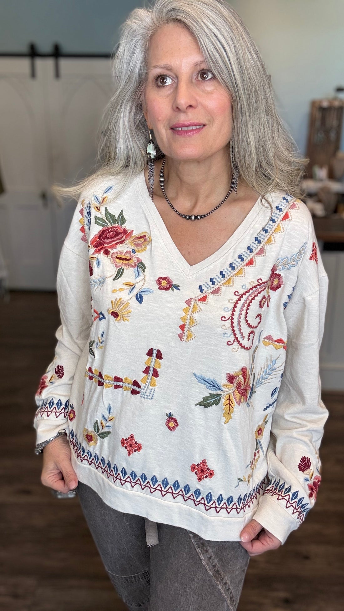 A woman with gray hair wearing a white long-sleeve V-neck top with colorful floral and paisley embroidery, paired with gray jeans, standing in an indoor setting.