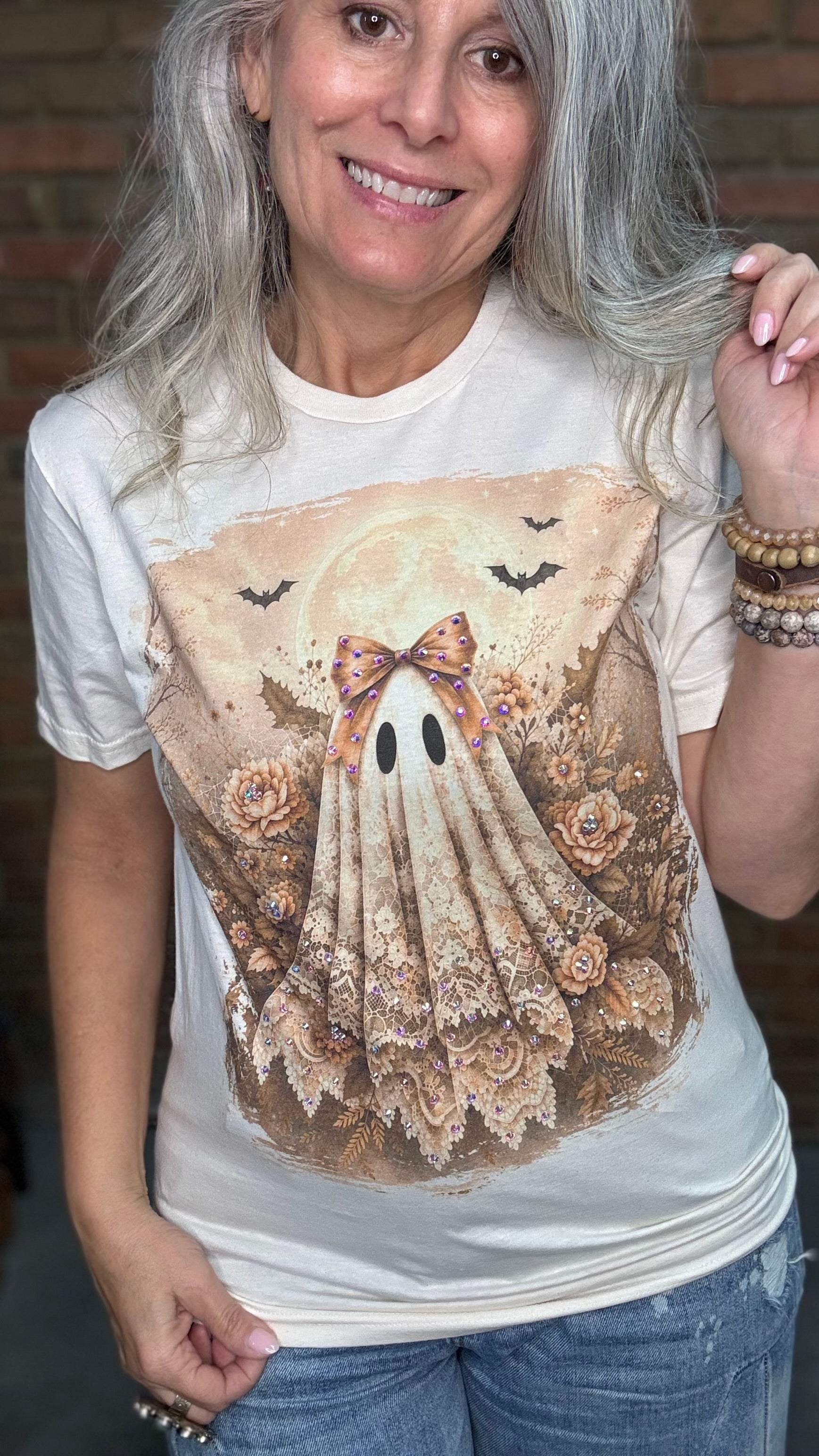 A woman with gray hair wearing a cream-colored graphic t-shirt featuring a vintage-style ghost illustration with lace, roses, and bats, paired with blue jeans.