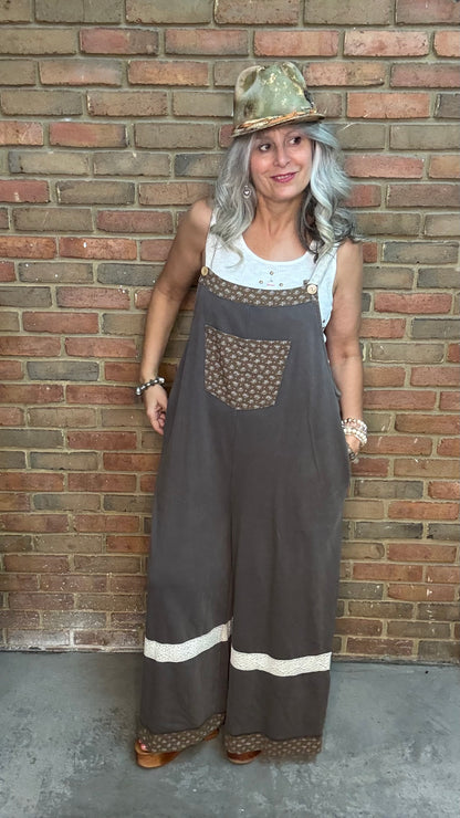 A woman with gray hair wearing a brown wide-leg overall jumpsuit with patterned details and a matching hat, posing in front of a brick wall.