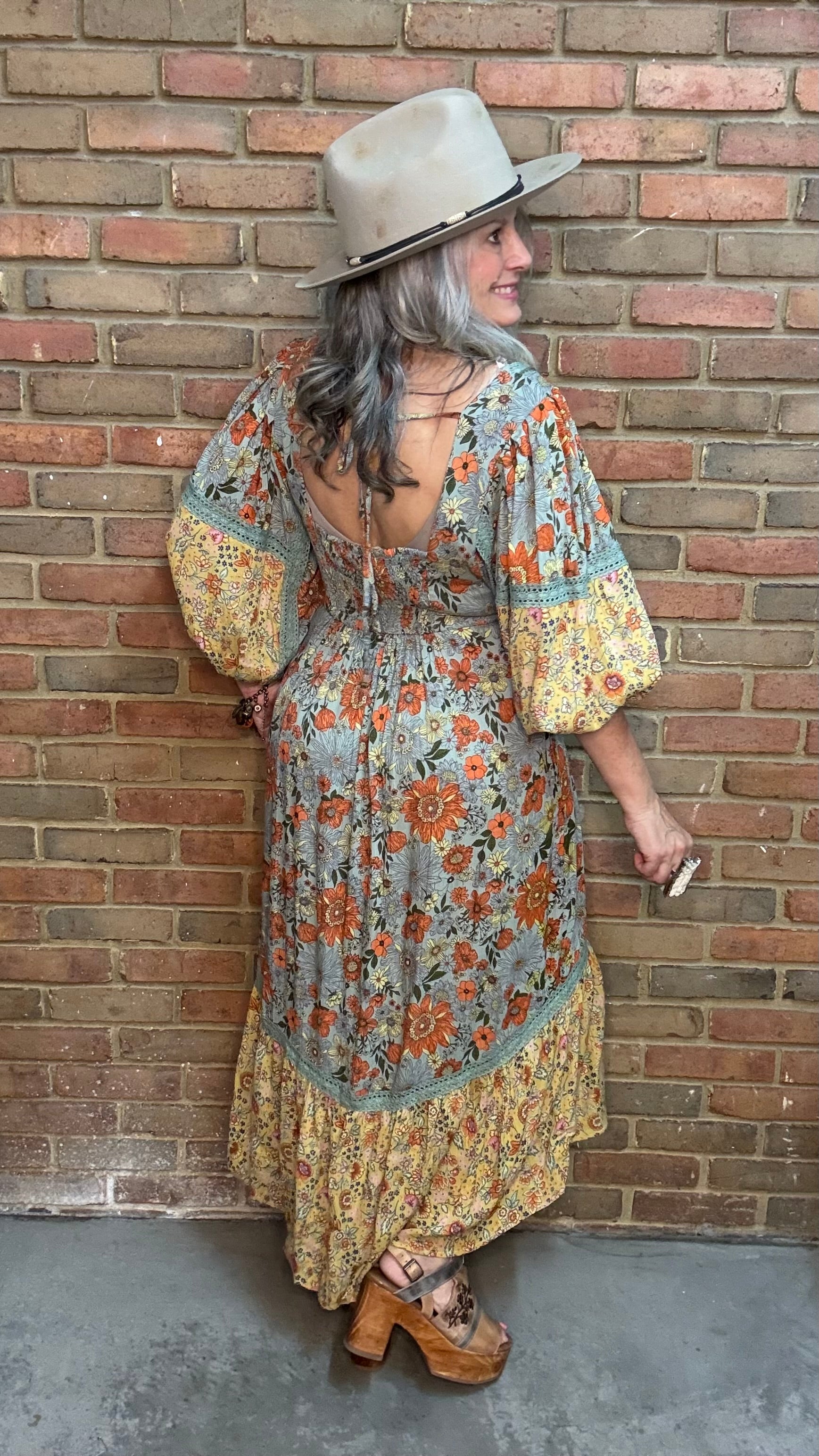 A woman stands against a brick wall wearing a bohemian-style midi dress, the  Long Sleeve V-Neck Hi-Lo Dress, with a blue-gray base and orange floral print, a wide-brimmed beige hat, and brown platform sandals.