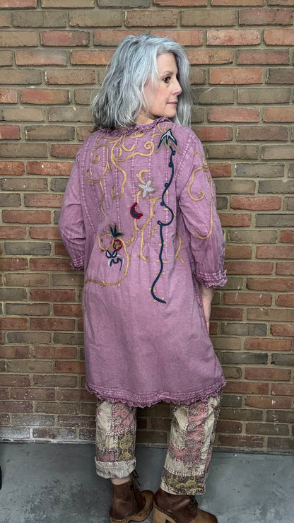 Person wearing a long, embroidered purple dress against a brick wall.