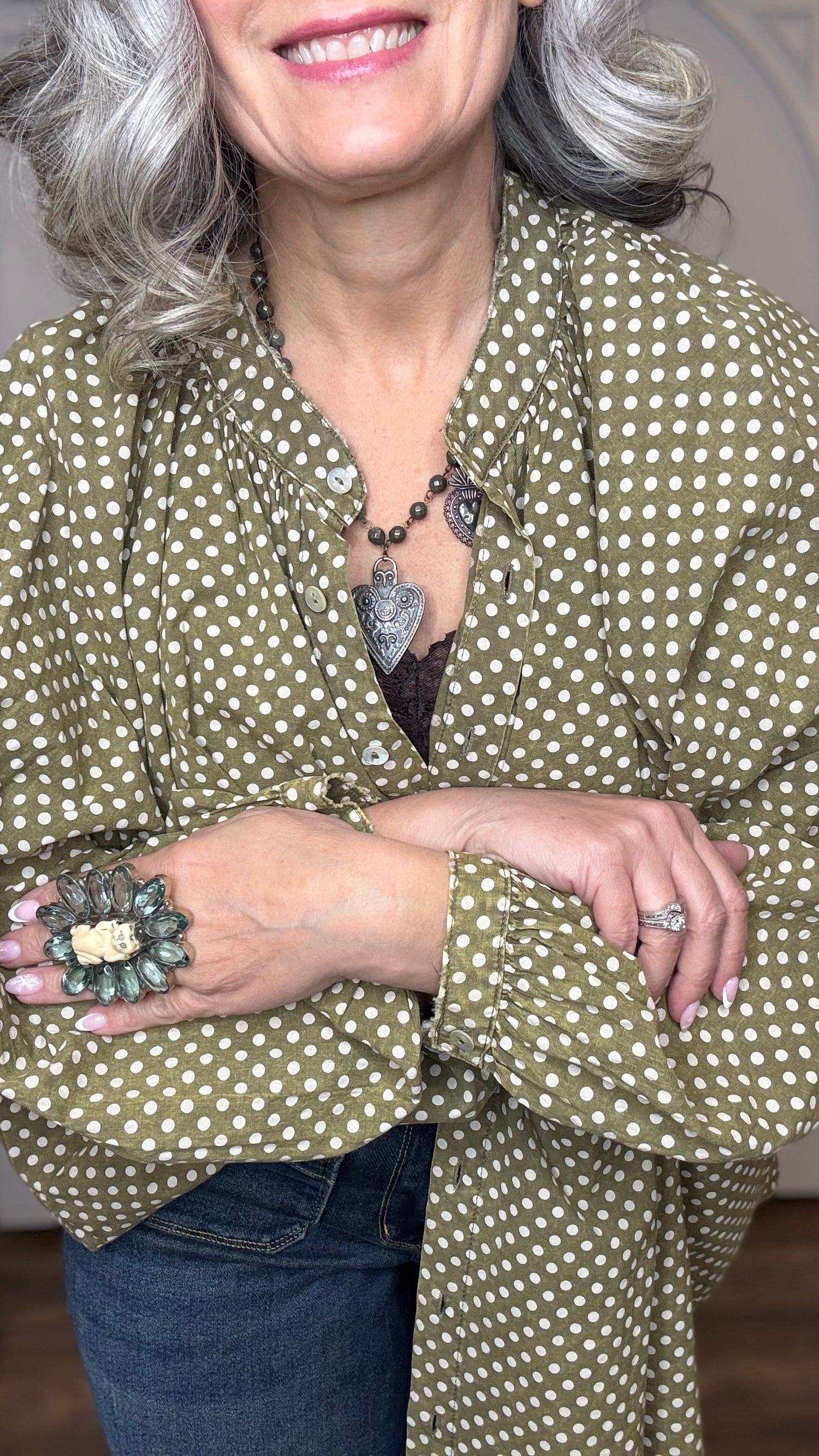 Woman wearing the Sweet Nothings Top-Olive Dot and Judy Blue Palazzo jeans paired with the Crystal Cat Ring and the Sacred Heart Charm Necklace standing against a white paneled wall.