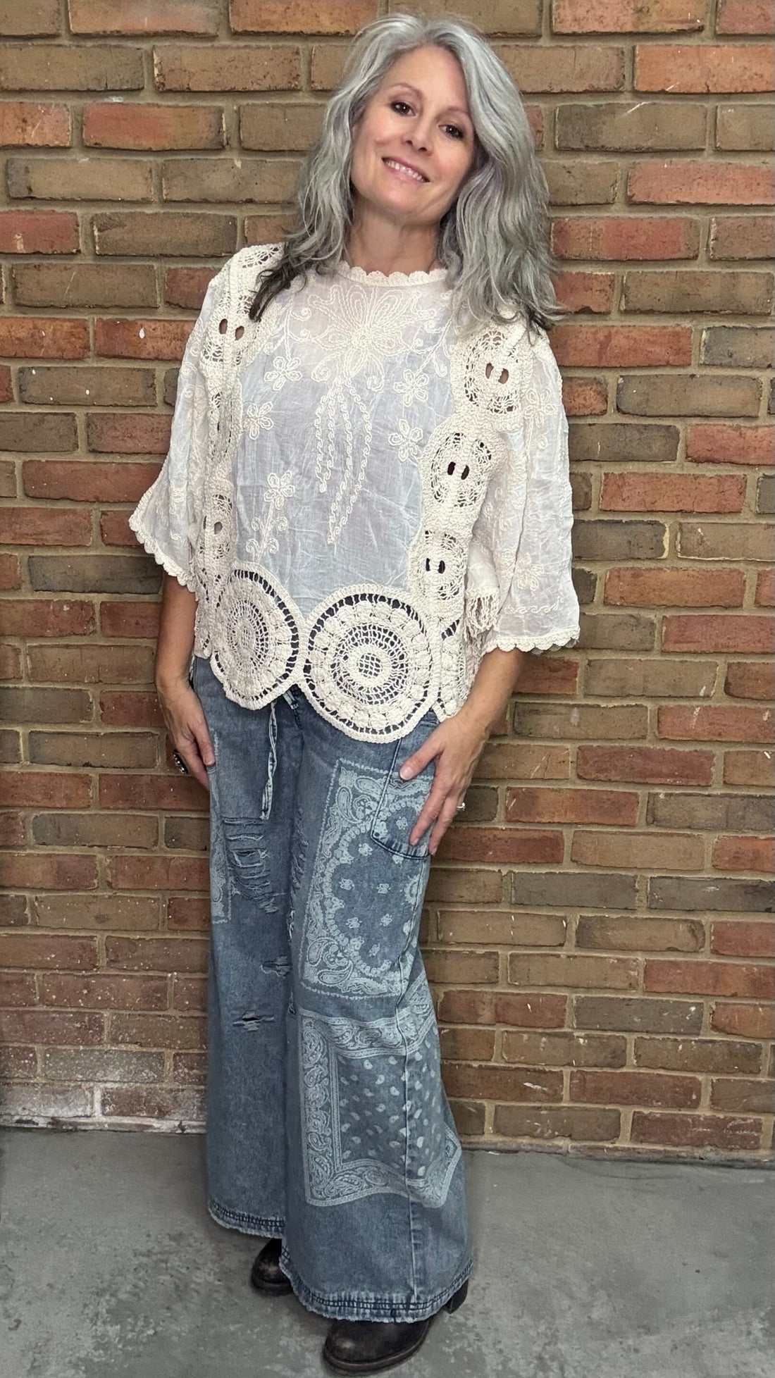 Woman wearing a white lace top and blue jeans standing against a brick wall.