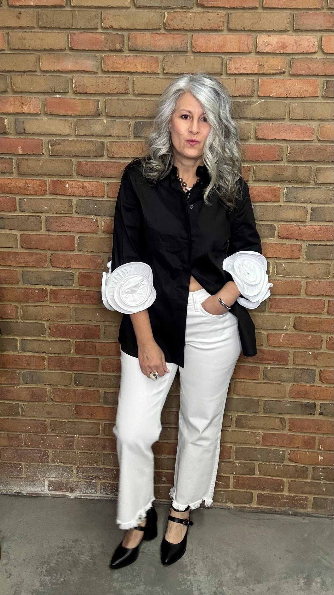 Woman wearing a black shirt with white ruffled sleeves and Mid Rise Uneven Hem Crop Jeans against a brick wall.