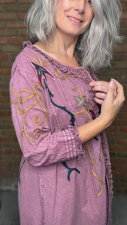Woman wearing a purple embroidered dress against a brick wall.