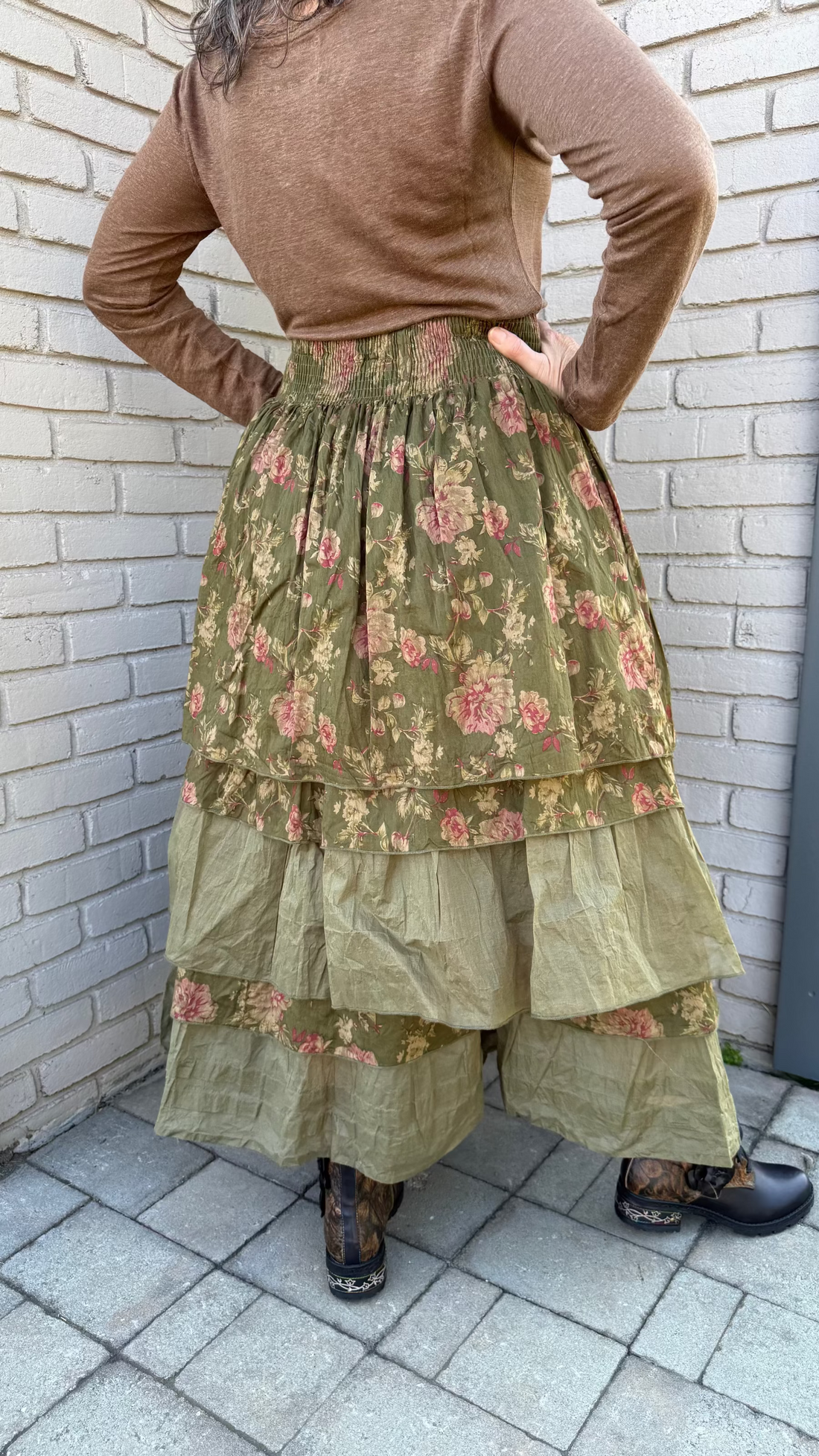 Person wearing a brown sweater and floral tiered skirt standing against a brick wall.