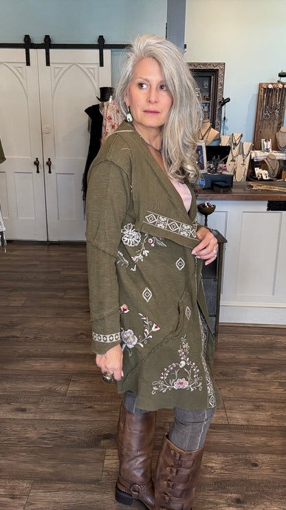 A woman with gray hair models an olive green embroidered cardigan with floral and geometric patterns, paired with brown leather boots in a rustic indoor boutique setting.