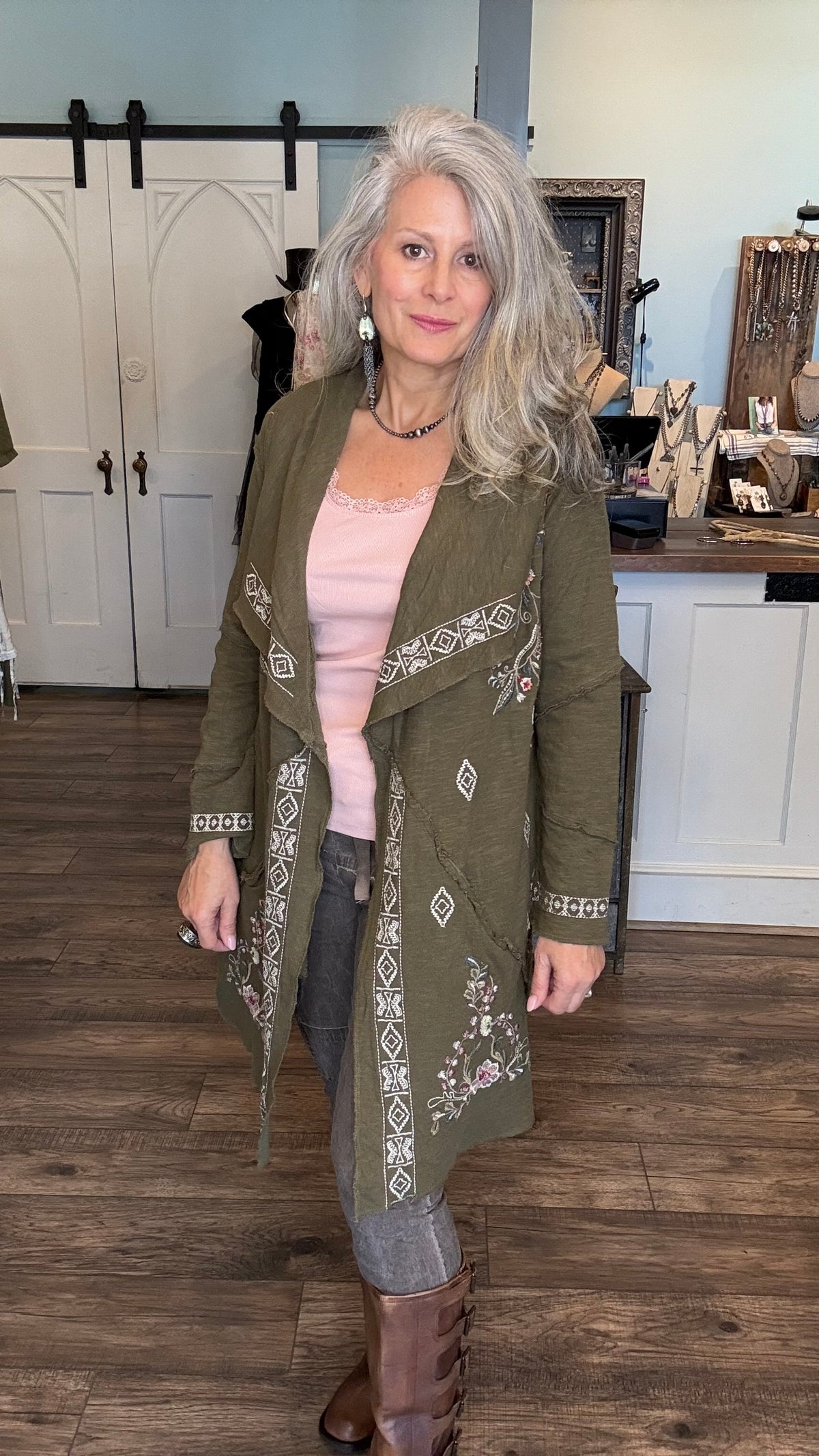 A woman with gray hair stands in a boutique wearing an olive green embroidered wrap coat over a light pink top, gray jeans, and brown boots.