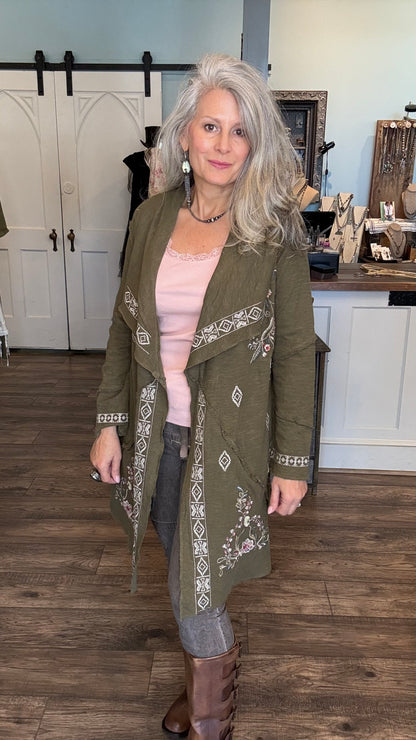 A woman with gray hair stands in a boutique wearing an olive green embroidered wrap coat over a light pink top, gray jeans, and brown boots.