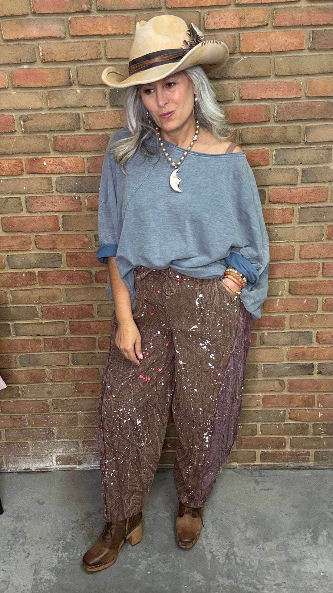 Woman wearing blue top and paint splatter pants in front of brick wall.