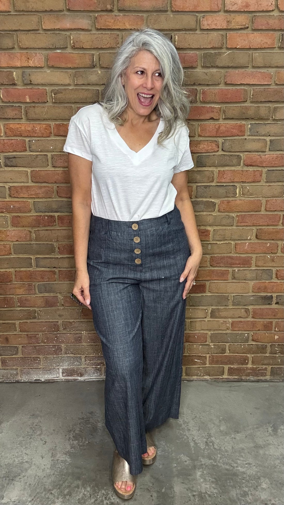 Woman wearing White Drop Shoulder V-Neck Top and Indigo Linen Button Front Pants standing against a brick wall.