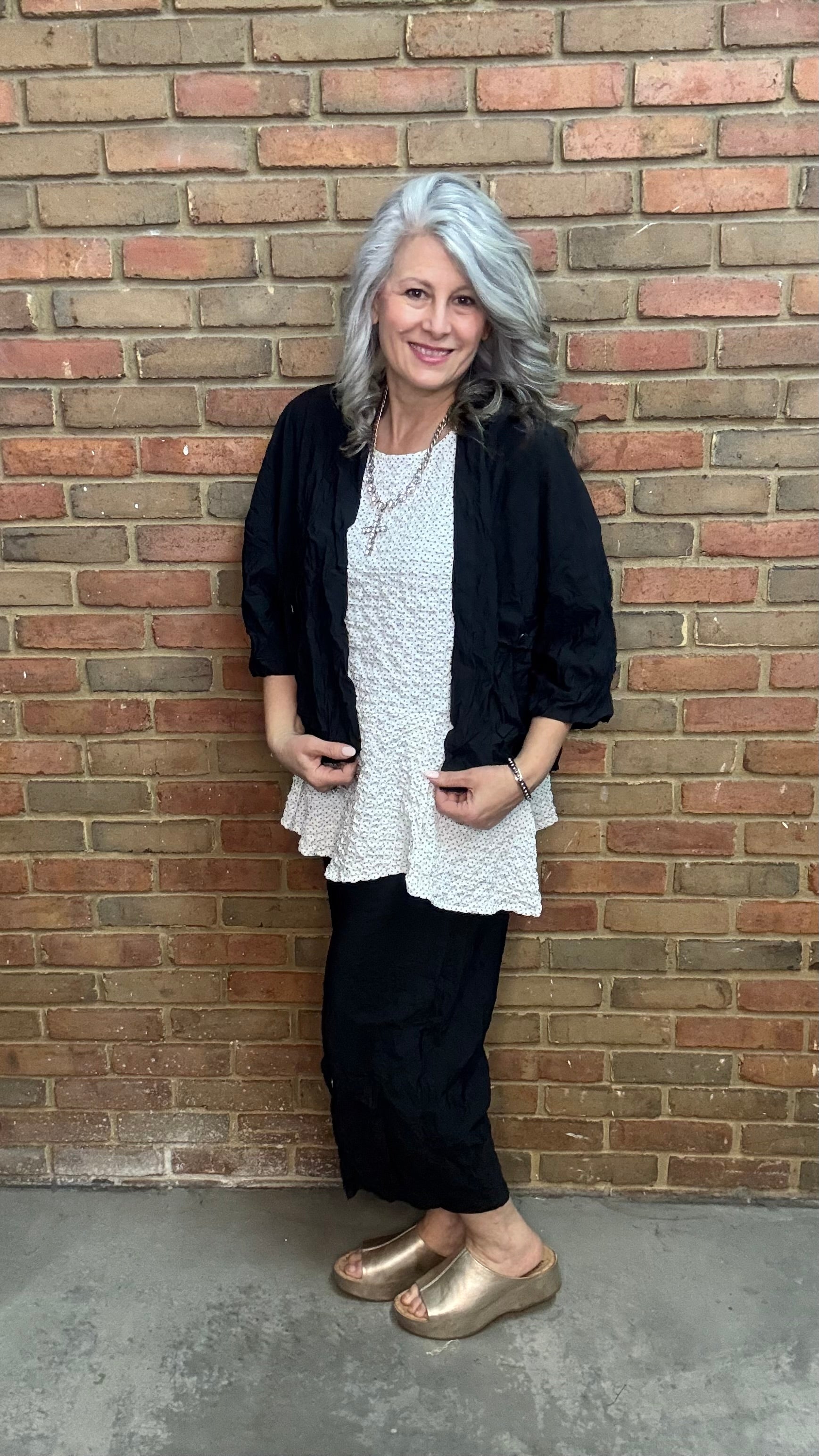 Woman standing against a brick wall wearing Francesca Jacket - Black, white top, and black pants.