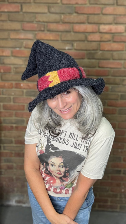 Person wearing a witch hat and t-shirt with a vintage-style graphic, standing against a brick wall.