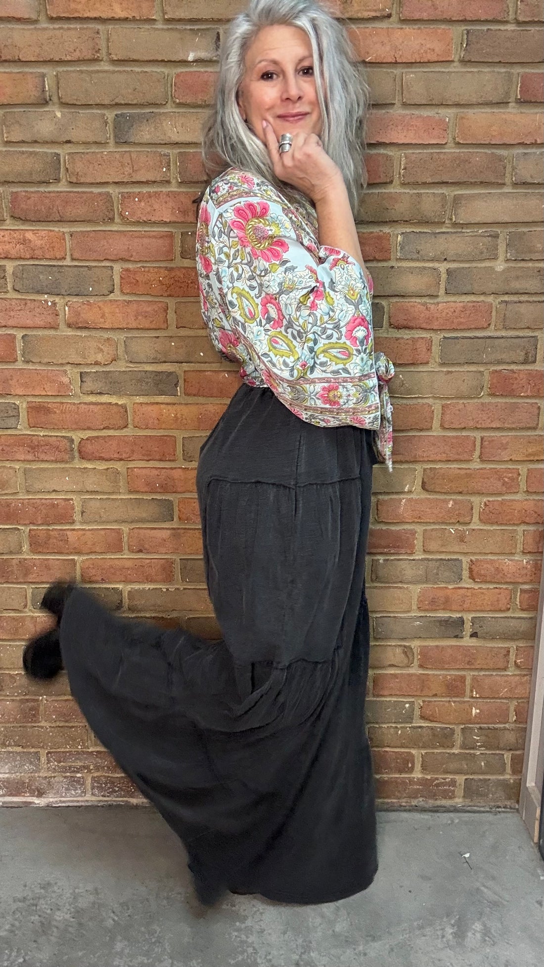 Woman wearing a floral Naomi Cross Back Top and Black Tiered Side Boho pants standing against a brick wall.