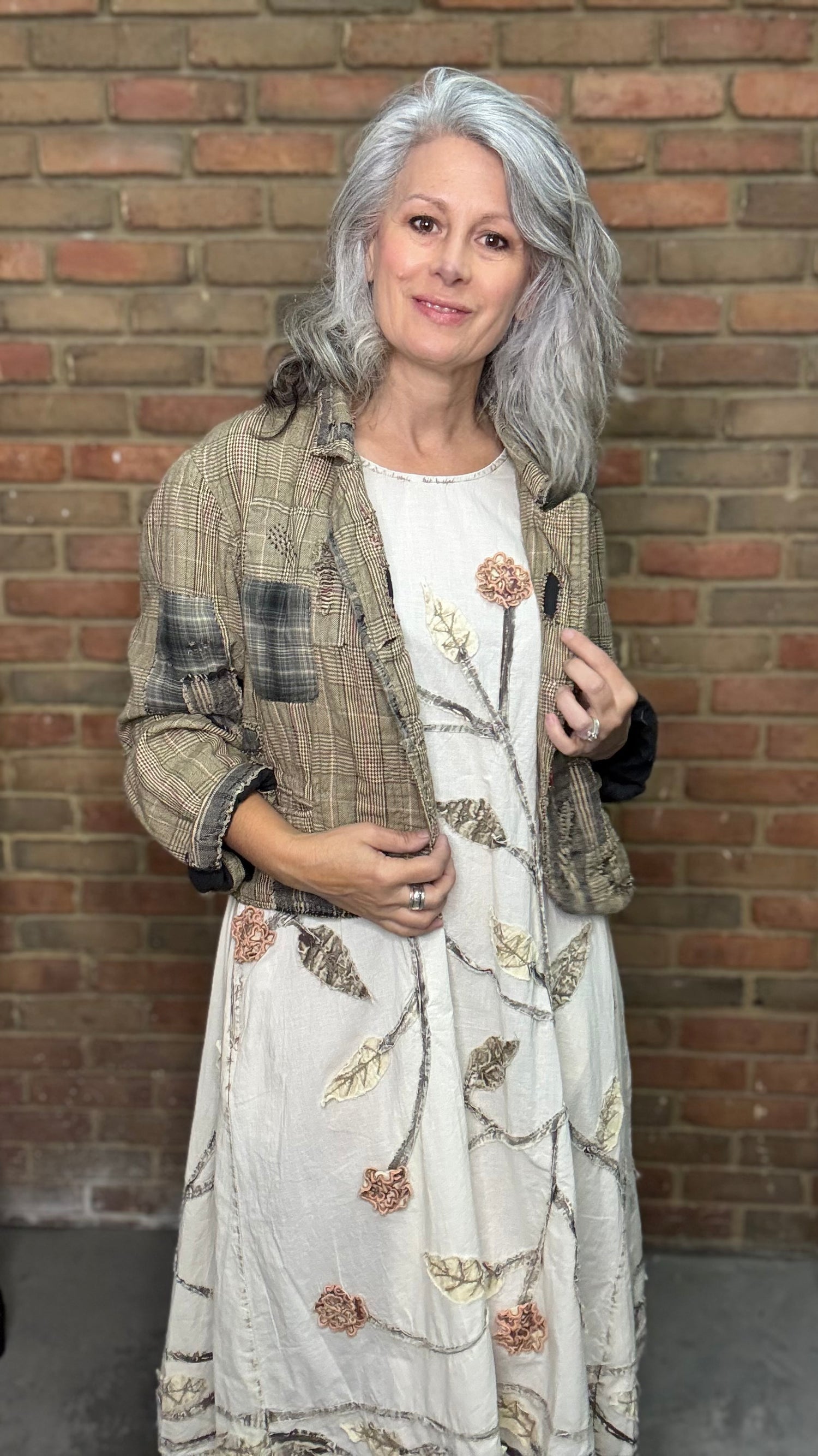 Woman wearing a patterned coat and floral dress against a brick wall.