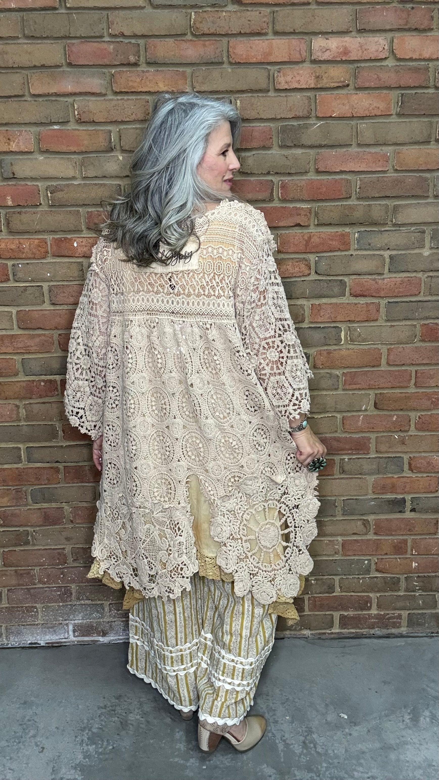 Woman wearing the crochet Forever Timeless Split Tails Jacket over a dress with striped pants standing against a brick wall.