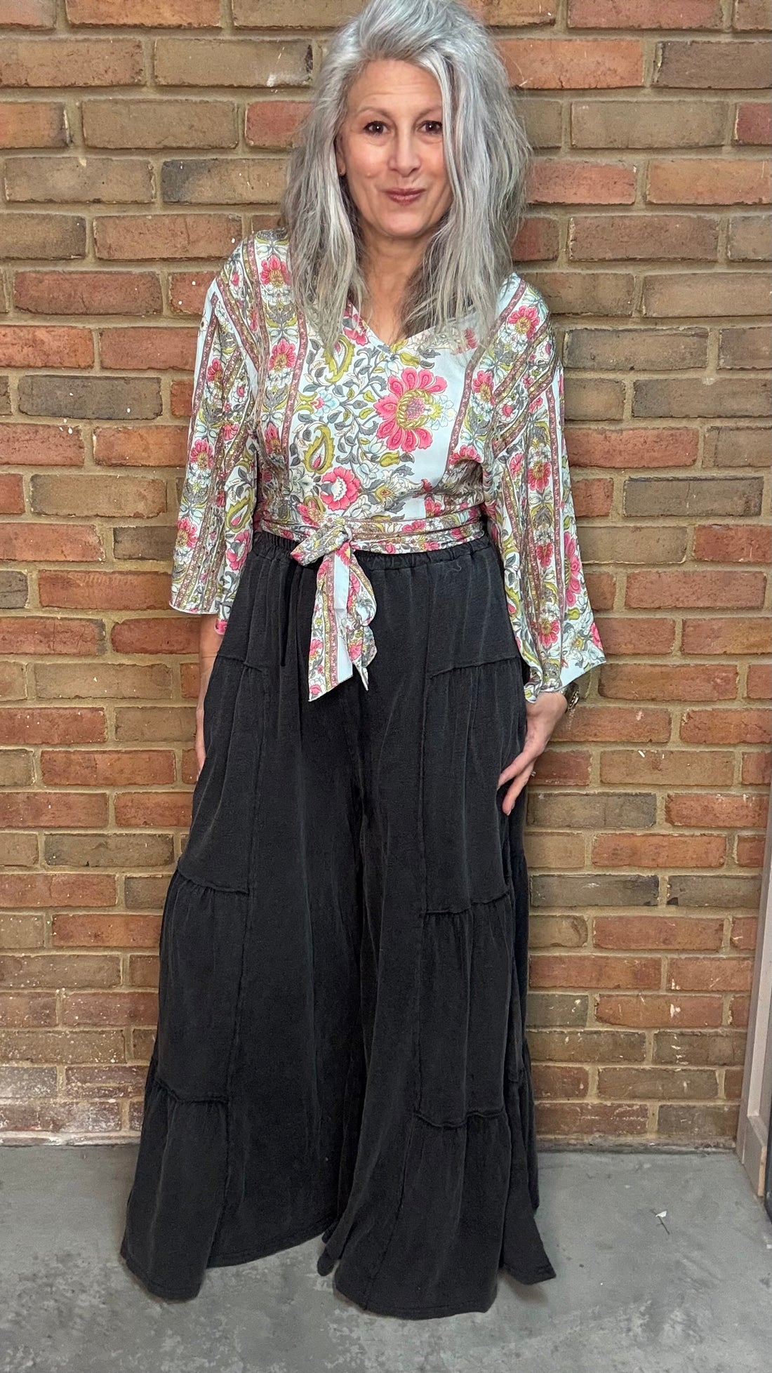 Woman wearing a floral Naomi Cross Back Top and Black Tiered Side Boho pants standing against a brick wall.