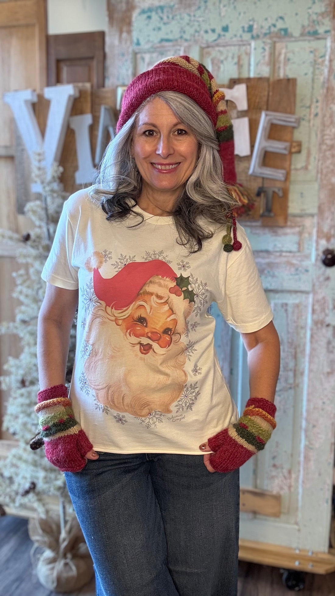 Person wearing a festive t-shirt with a Santa Claus design, colorful hat, and gloves in a home setting.