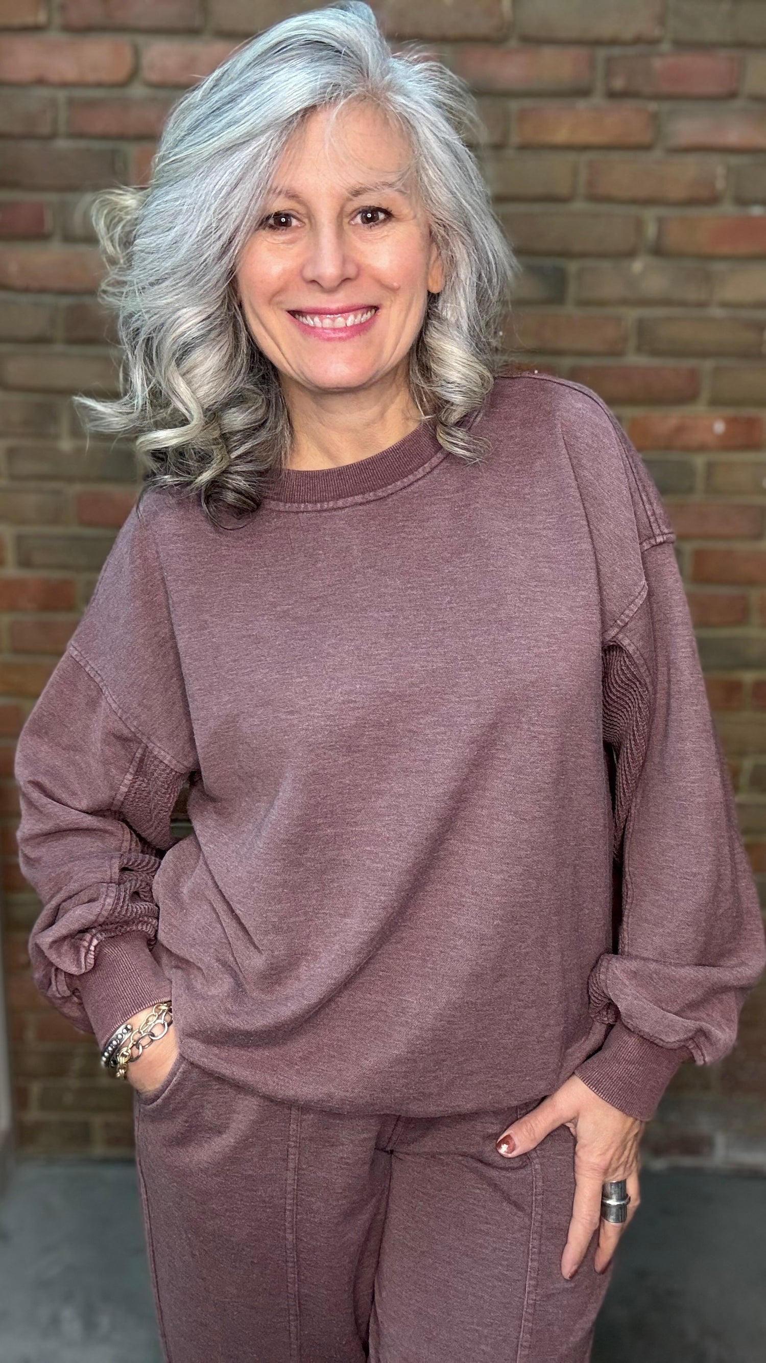 Woman wearing a mauve sweatshirt and pants standing in front of a brick wall.