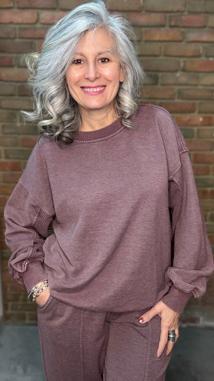 Woman wearing a mauve sweatshirt and pants standing in front of a brick wall.
