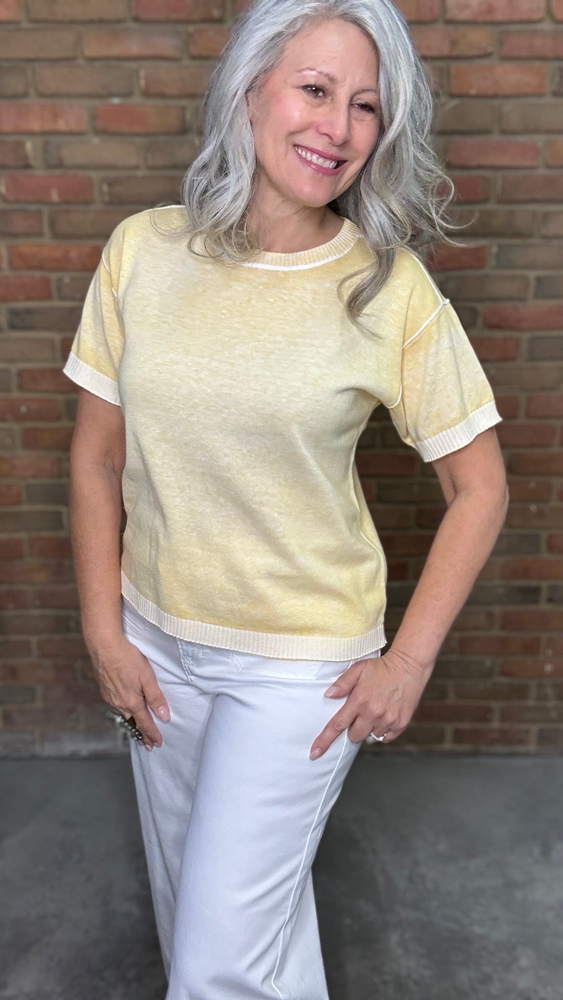 Woman wearing the Sunshine Short Sleeve Sweater, a light yellow sweater and white pants standing against a brick wall.
