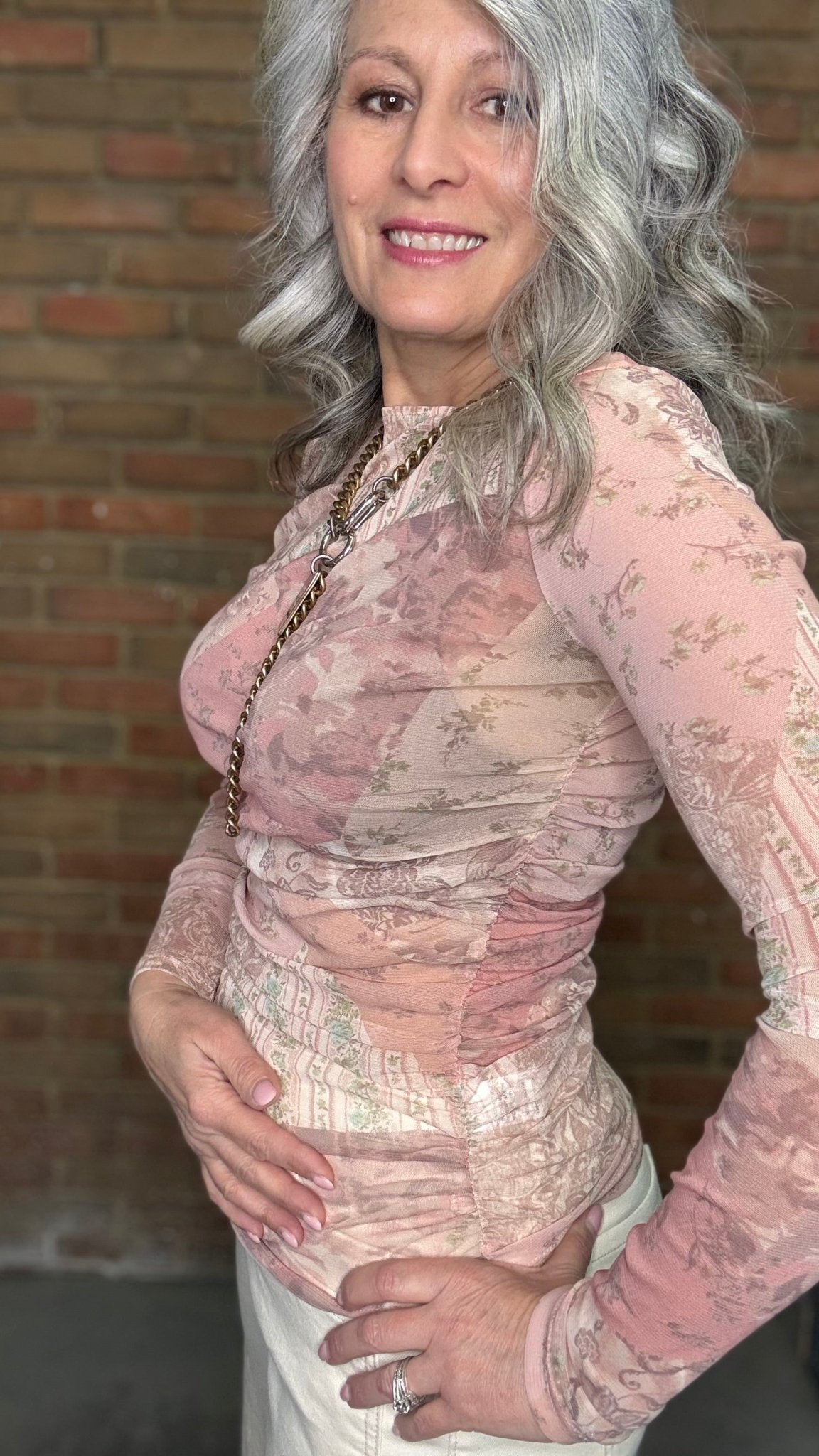 Woman wearing Mix Print Long Sleeve Mesh Top and white pants standing against a brick wall.