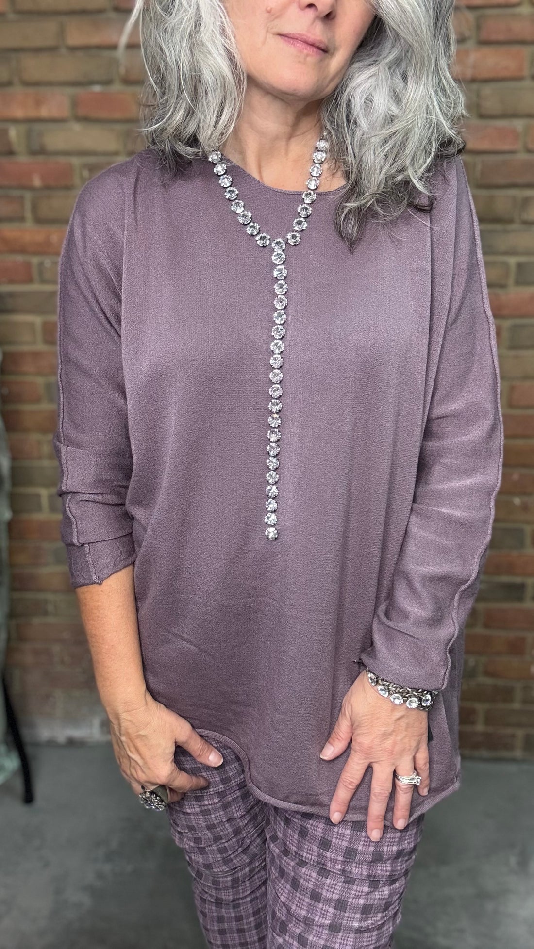 Person wearing a long-sleeve purple top with a decorative necklace against a brick wall.