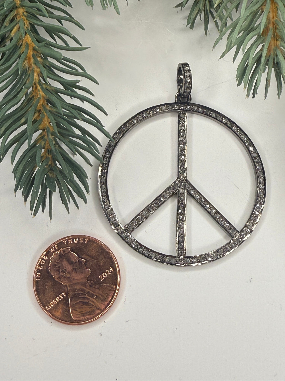 Silver peace symbol pendant next to a penny on a white background with greenery