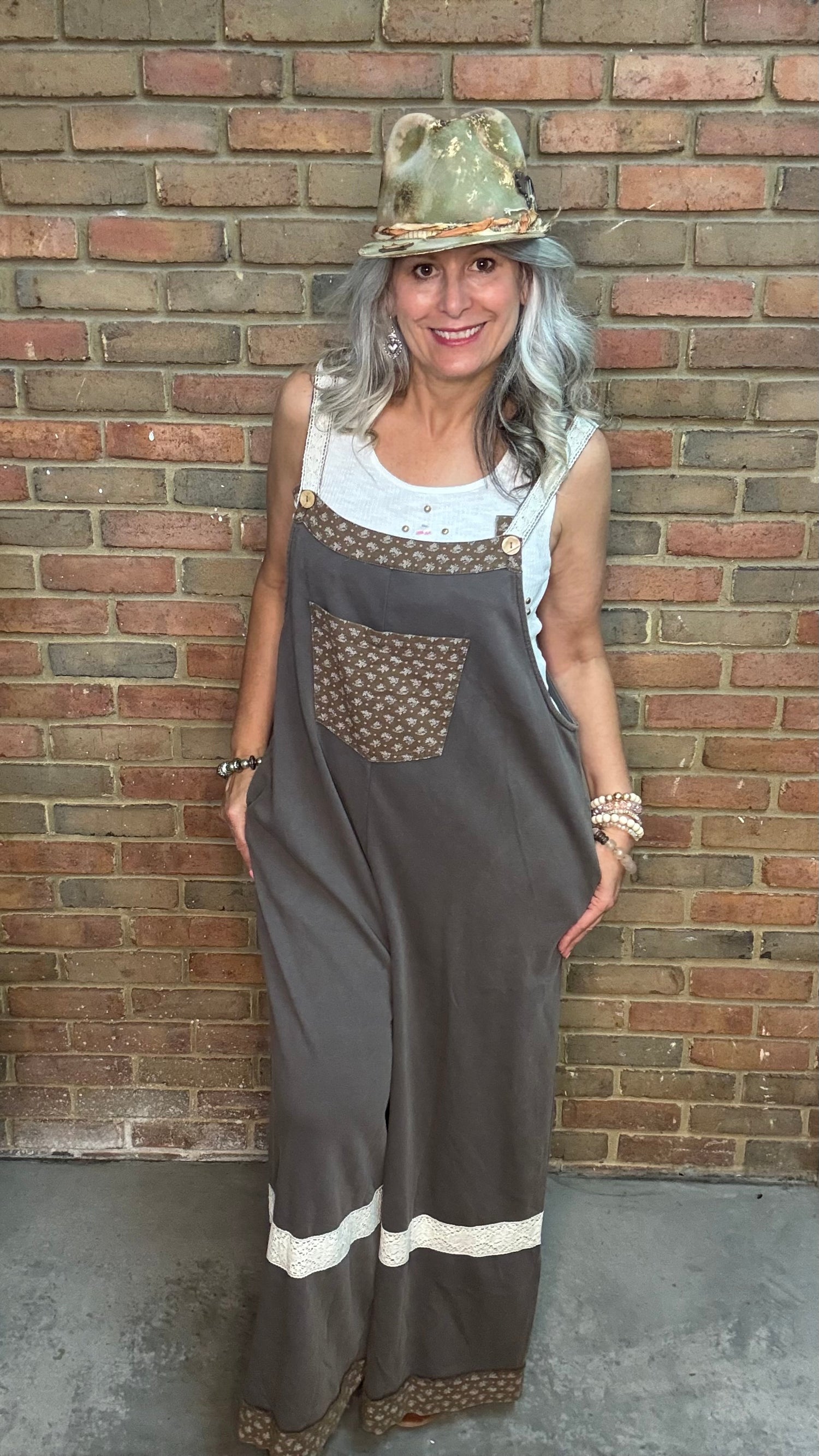 A woman with gray hair wearing a brown wide-leg jumpsuit with patterned chest pocket and white stripe details, a white tank top, and a gold hat, standing against a brick wall.