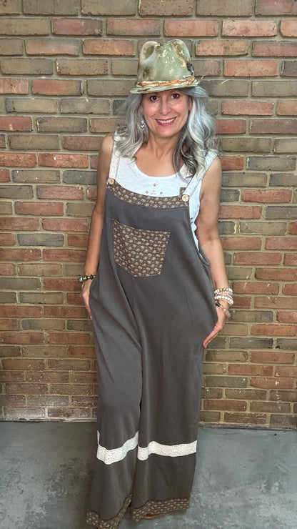 A woman with gray hair wearing a brown wide-leg jumpsuit with patterned chest pocket and white stripe details, a white tank top, and a gold hat, standing against a brick wall.