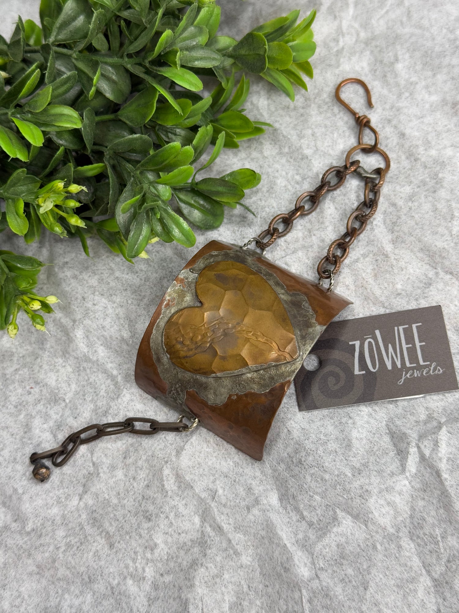 Heart-shaped design on a half cuff with chain on a textured surface next to a plant
