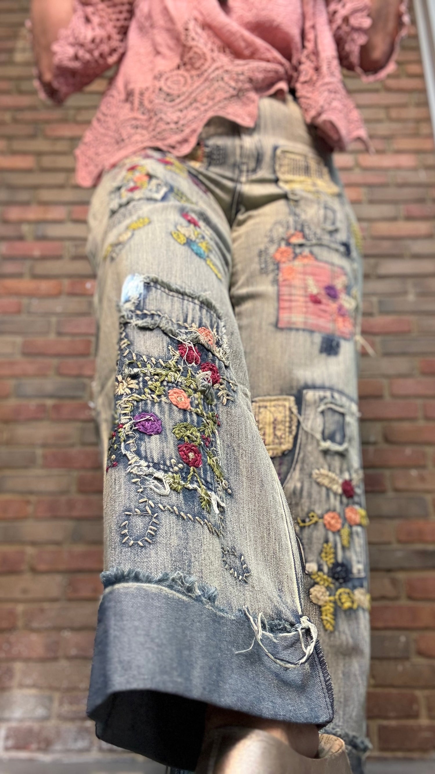 A close-up of a person wearing distressed denim culottes with colorful floral embroidery, patchwork details, and frayed hems, paired with a pink lace top, set against a brick wall background.