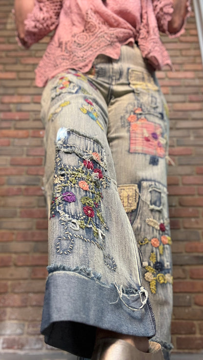 A close-up of a person wearing distressed denim culottes with colorful floral embroidery, patchwork details, and frayed hems, paired with a pink lace top, set against a brick wall background.