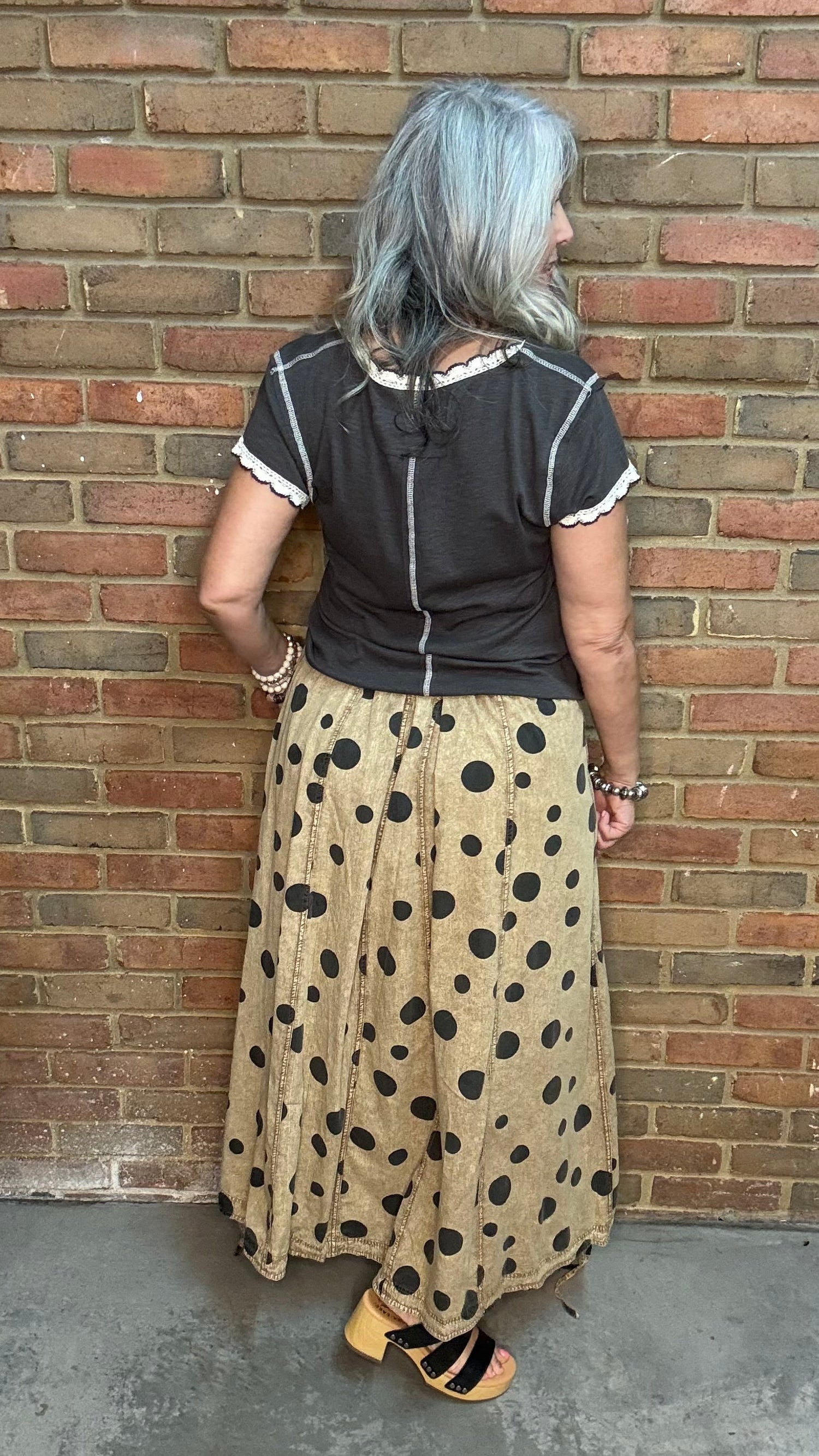 A woman with gray hair is shown from the back, wearing a black short-sleeve top with white trim and a beige maxi skirt with black polka dots, standing against a brick wall.