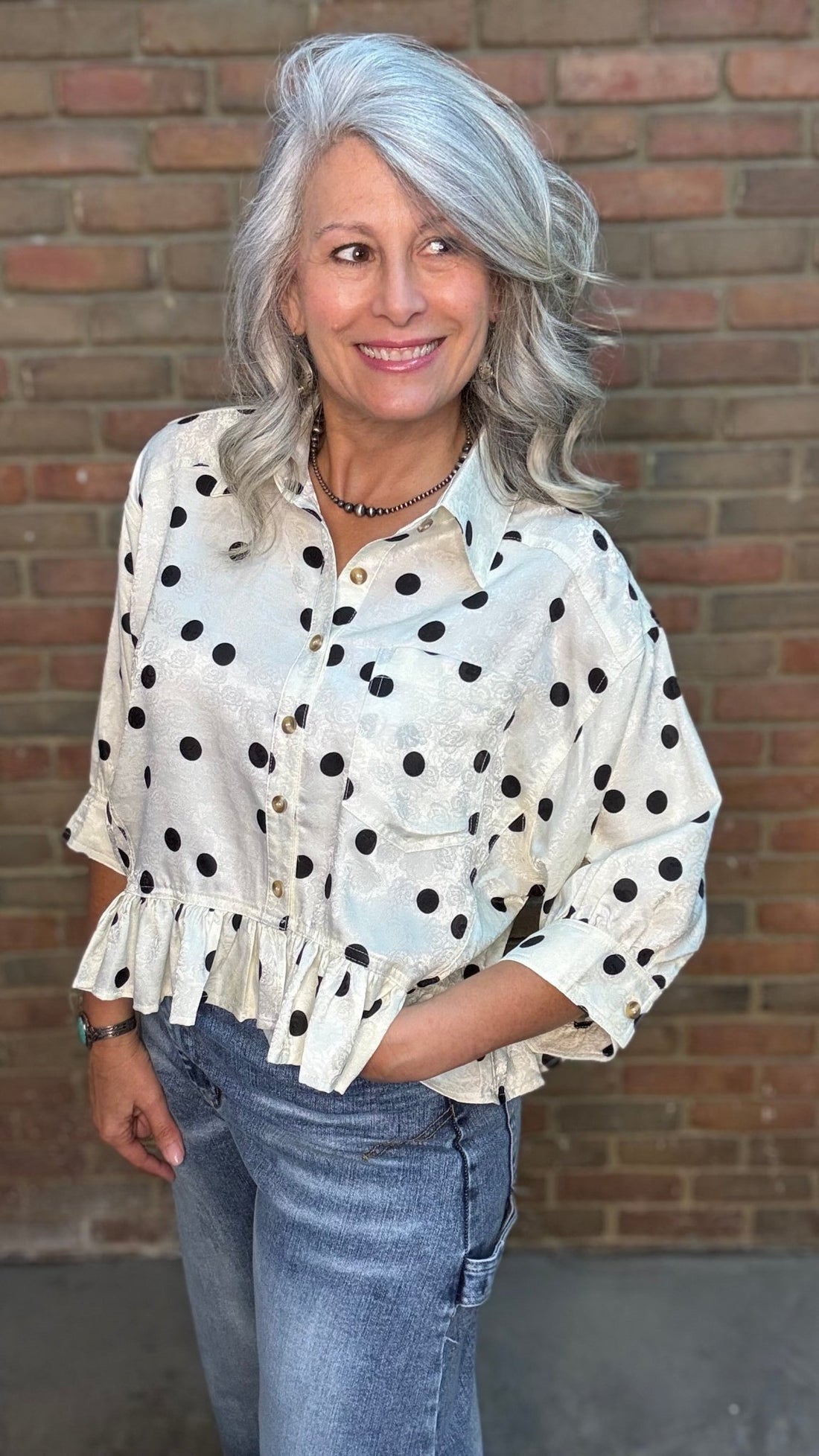 Woman wearing the Polka Dot Ruffle Hem Shirt and jeans against a brick wall.