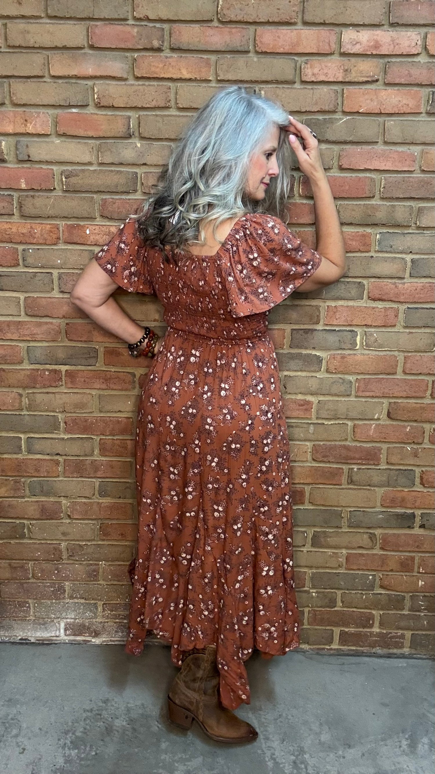 A woman with gray hair stands against a brick wall wearing a rust-brown bohemian-style midi dress with white floral prints, the "Short Sleeve Embroidered Midi Dress" and brown cowboy boots.