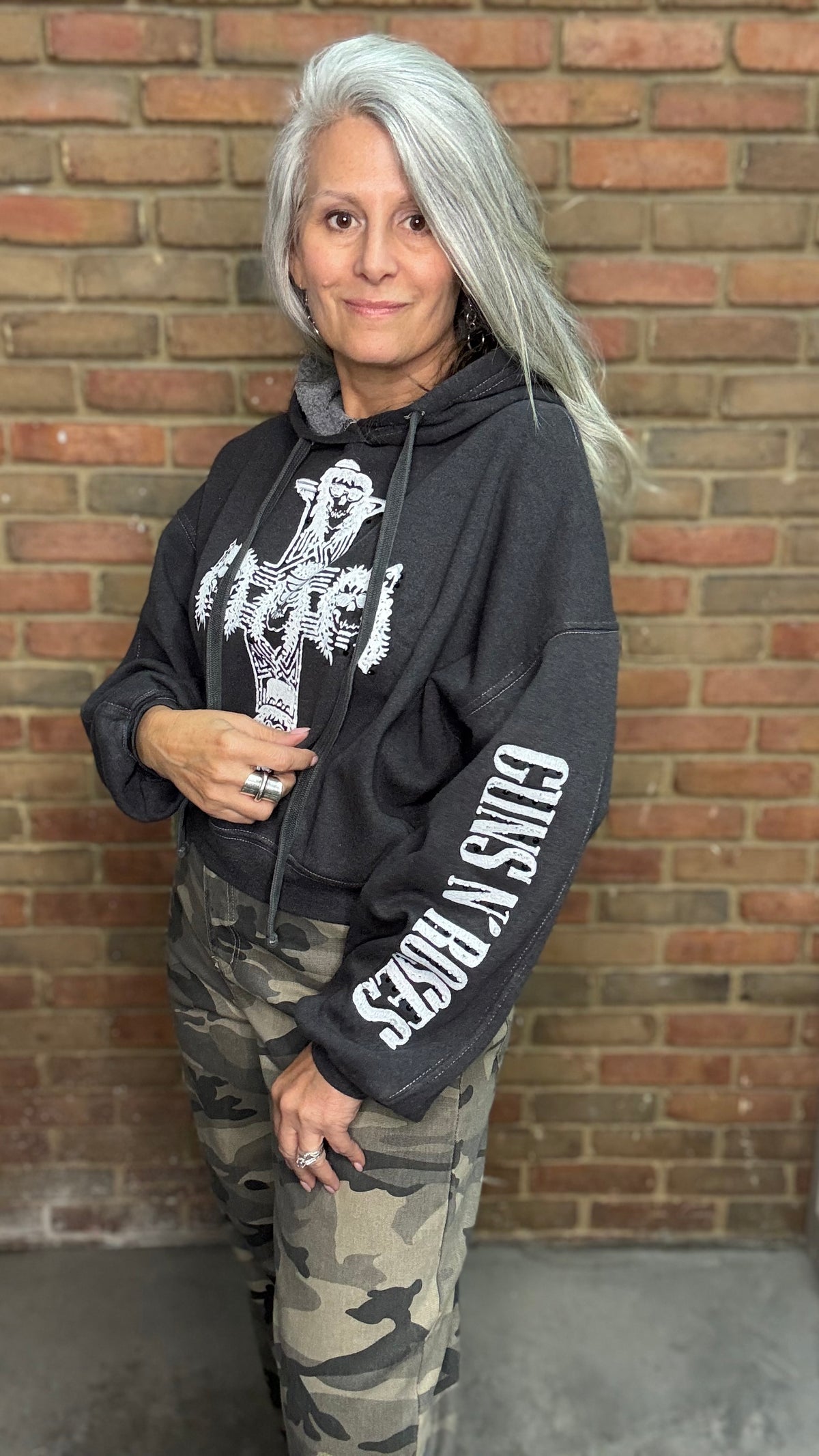 A woman with long silver hair stands in front of a brick wall wearing a black Guns N&