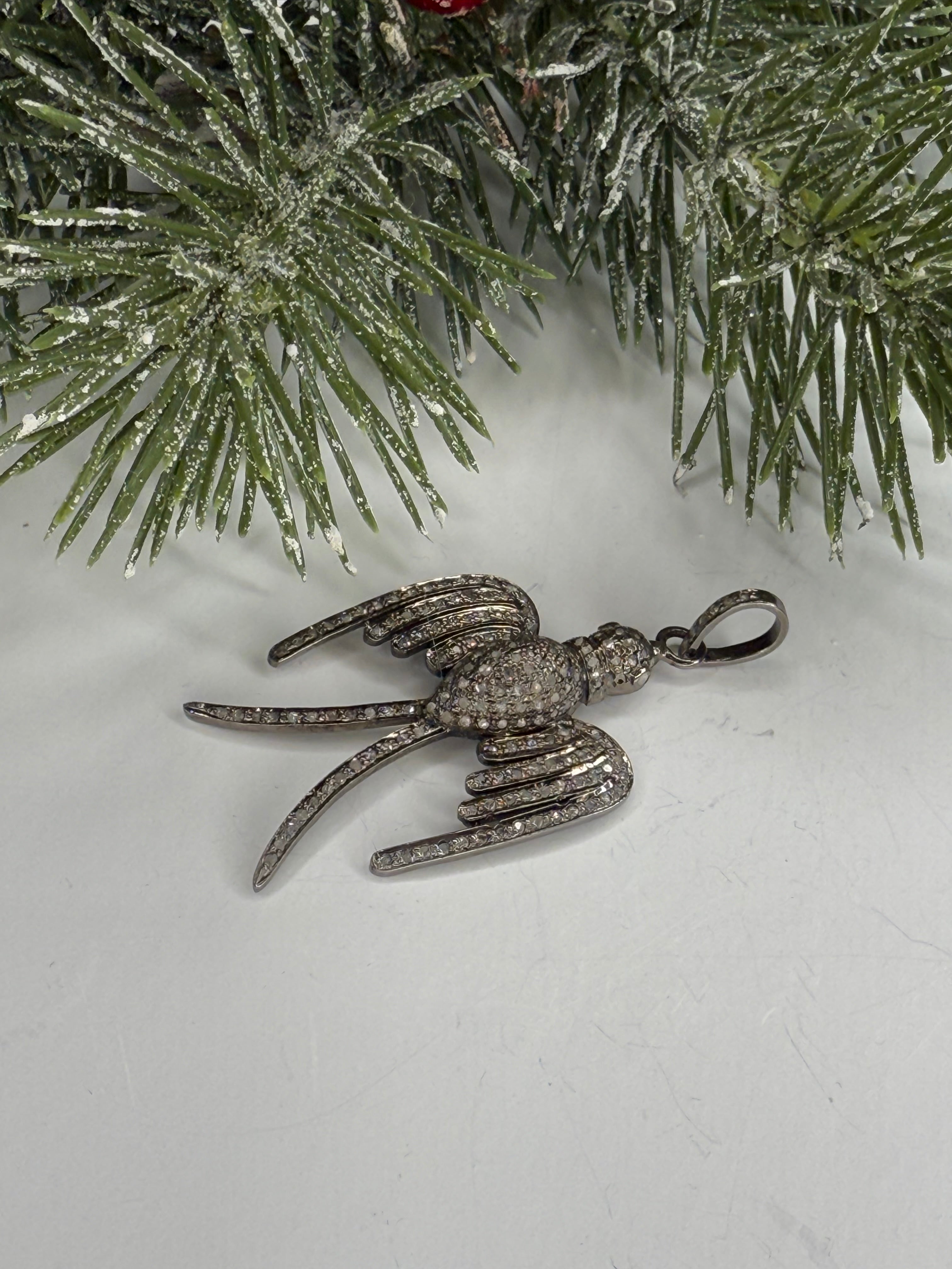 Bird shaped pendant in pave diamonds on a white surface with a Christmas tree branch in the background.